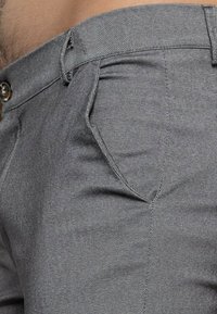 Pantalon en denim gris avec un motif chevrons subtil, poche avant et fermeture par bouton. Le tissu semble lisse et légèrement texturé.