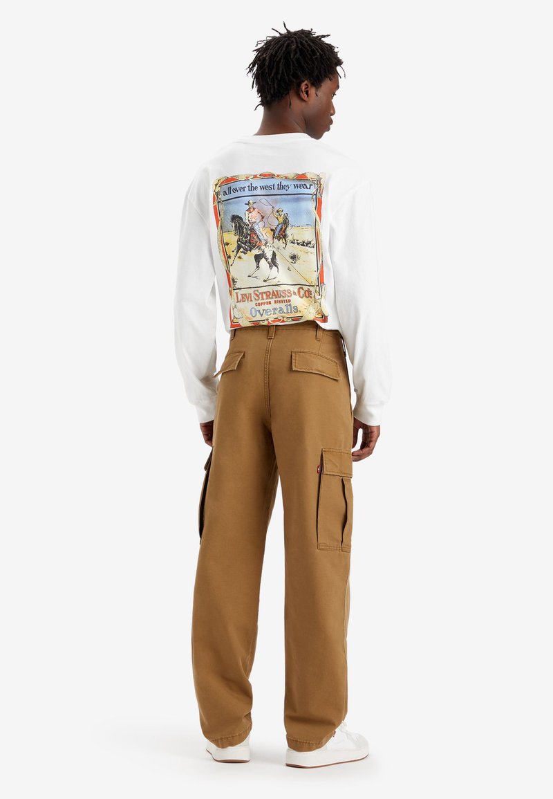 Camisa blanca de manga larga con un gráfico vintage en la parte posterior. Pantalones cargo marrones con múltiples bolsillos y un corte relajado. Zapatillas blancas.