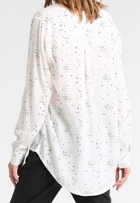 Camisa blanca de manga larga con un patrón de puntos negros, que presenta un dobladillo redondeado, puños abotonados y detalle de canesú en la parte trasera.