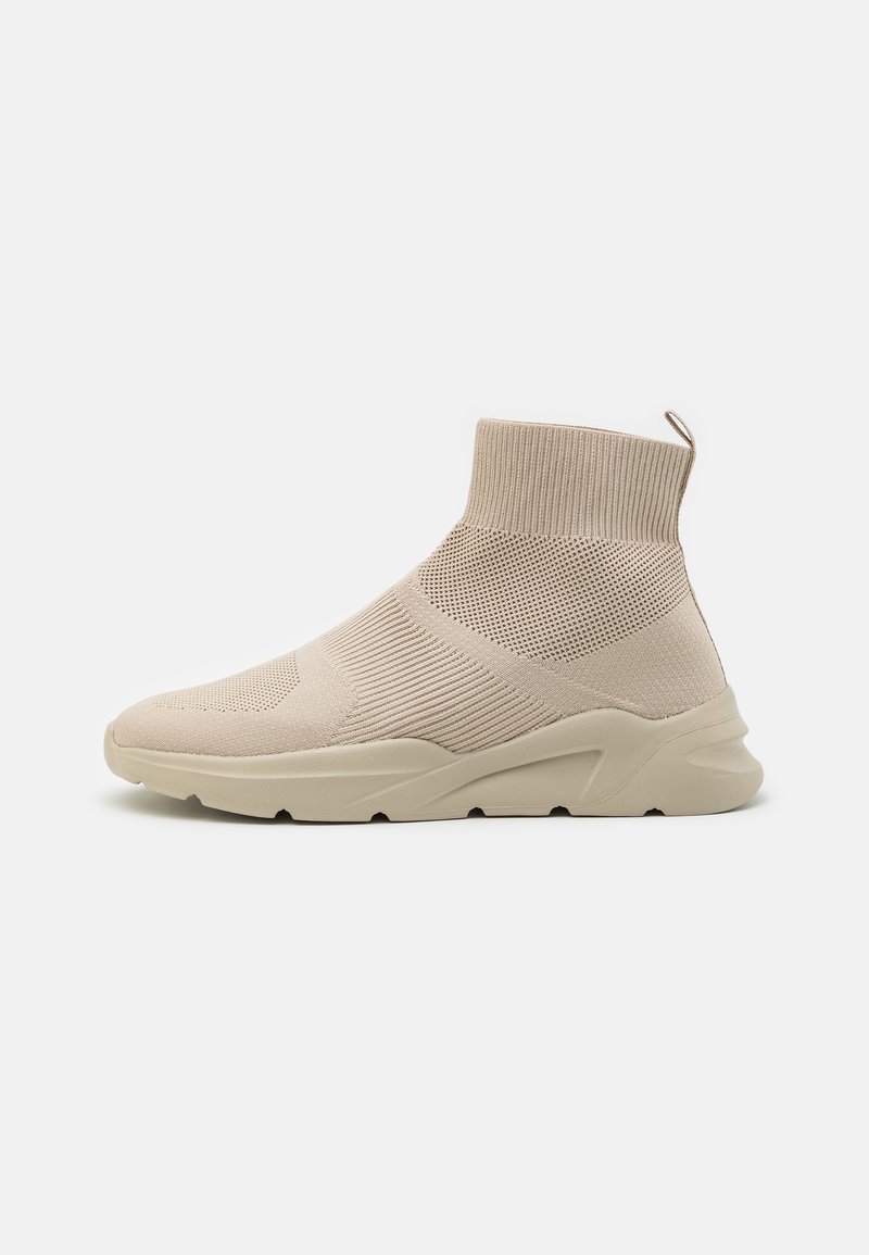 Beige strikket slip-on joggesko med en sokklignende overdel, teksturerte paneler og en tykk, polstret såle. Har en trekksløyfe på hælen.