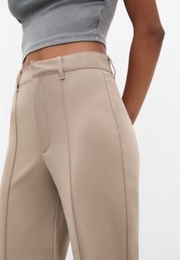 Pantalon beige sur mesure avec une texture lisse, une coupe droite, et des poches avant. Détails de coutures le long des côtés.