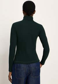 Person mit geflochtenem Haar, der einen dunkelgrünen Rippstrickpullover mit Stehkragen und blaue Jeans trägt, von hinten gezeigt.