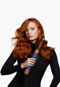 Spazzola per asciugacapelli con cilindro, dettagli arancioni, corpo blu scuro e setole testurizzate; utente che la tiene con i lunghi capelli rossi acconciati.
