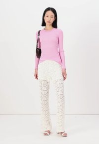 Tricou lung cu mâneci, roz, cu decolteu zimțat; pantaloni albi din dantelă cu model floral; model purtând sandale albe și ținând o geantă neagră.
