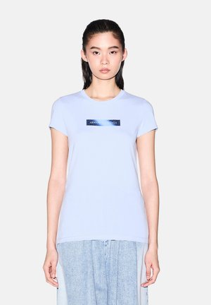 Joven mujer con cabello oscuro y liso lleva una camiseta azul claro de Armani Exchange y una falda vaquera clara, de pie frente a un fondo sencillo.