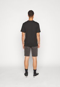 Hombre de pie de espaldas vistiendo una camiseta negra, pantalones cortos de mezclilla negros desvanecidos, calcetines negros y zapatillas negras de corte bajo sobre fondo blanco.