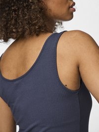 Gros plan sur une personne aux cheveux bouclés portant un haut sans manches côtelé bleu marine, montrant son épaule et son profil latéral sur un fond blanc.