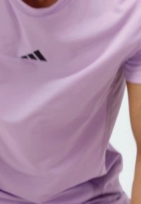 Camiseta atlética de color púrpura claro hecha de una tela suave, con un logo negro de Adidas en el pecho. El diseño incluye mangas cortas y un ajuste relajado.