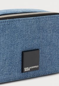 Modrá denimová taška so strieborným zipsom. Obsahuje čierny obdĺžnikový logo nášivku s bielym textom "KARL LAGERFELD JEANS." Textúrovaný povrch.
