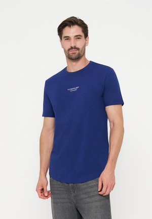Homme aux cheveux courts et foncés avec une barbe, portant un t-shirt bleu avec le texte "AUTHENTIC DEPT" et un jean gris, debout devant un fond blanc.