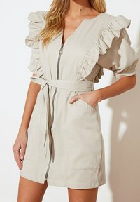 Béžové krátké šaty s volánkovými rukávy, zipem vpředu a páskem v pase. Obsahují boční kapsy a volný střih. Měkká, lehká tkanina.