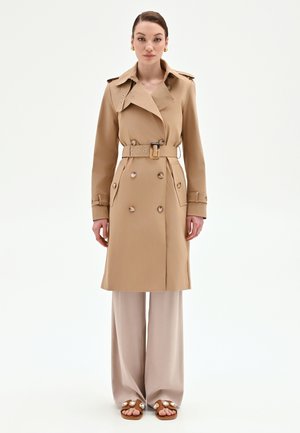 Donna in piedi di fronte, indossa un trench beige con cintura, pantaloni a gamba larga e sandali marroni con grandi decorazioni.