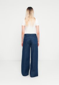 Vrouw met lang blond haar, gekleed in een wit T-shirt met korte mouwen en wijde blauwe spijkerbroeken, staande tegenover een witte muur.