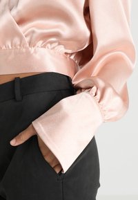 Personne portant un chemisier en satin rose pâle brillant avec de larges poignets, main dans la poche d'un pantalon noir taille haute, sur un fond uni.