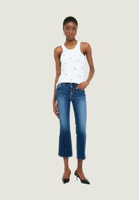 Top bianco senza maniche con ricami floreali, abbinato a jeans in denim blu a vita alta con bottoni e un leggero sbiadimento, completato da tacchi neri.