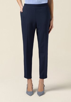 Mujer con pantalones tobilleros azul marino de corte ajustado y camisa azul claro, con una mano en el bolsillo y tacones gris de punta afilada sobre un fondo claro.