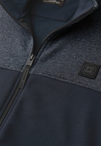 Gros plan sur une veste zippée bleu marine et gris foncé avec un tissu supérieur texturé, un patch de marque noir et une tirette de fermeture éclair portant l'inscription « SEEK ADVENTURE ».