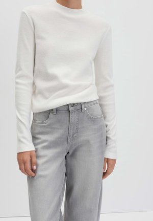 Personne portant une chemise blanche à manches longues rentrée dans un jean gris clair taille haute, debout devant un fond uni.