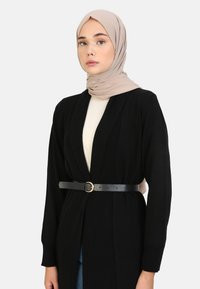 Zwarte cardigan met een riem en details van goudkleurige hardware. Gedragen over een crèmekleurige top met lange mouwen en een open voorkant.