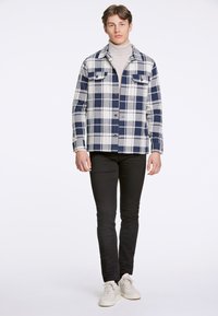 Giacca a camicia a quadri in blu navy e bianco, con chiusura a bottoni, due tasche sul petto e una vestibilità comoda, abbinata a pantaloni neri slim e sneakers.