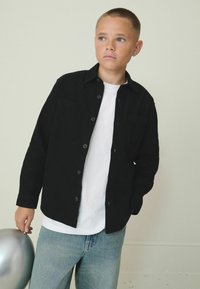 Schwarze Hemdjacke mit zwei Brusttaschen, über einem weißen T-Shirt getragen, kombiniert mit hellblauen Jeans, gehalten mit einem silbernen Ballon.