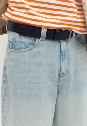 Gros plan sur un jean en denim bleu clair avec une ceinture en cuir noire et une chemise rayée orange et blanche rentrée.