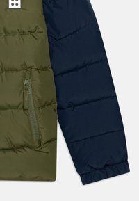 Detailný záber rukáva dvojfarebnej prešívanej bundy v olivovo zelenej a námorníckej modrej farbe, s vreckom na zips a elastickým manžetovým zakončením.