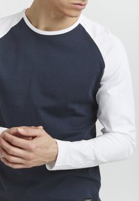 Sötétkék, hosszú ujjú póló fehér raglán ujjakkal, kerek nyakkivágással és puha pamut anyagból készült. Az alján egy címke található.