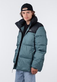 Veste rembourrée avec un corps teal et une section épaule noire. Présente une fermeture éclair, un col haut et des coutures visibles. Portée par-dessus une chemise noire.