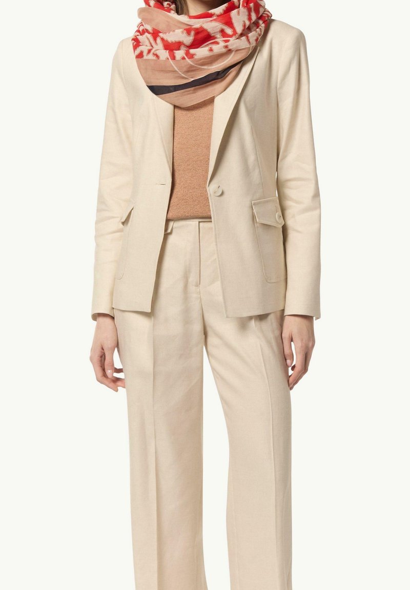 Femme portant un costume beige avec une veste et un pantalon sur mesure, un pull marron clair et une écharpe rouge et beige à motifs autour du cou.