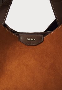 Brun semsket veske med en teksturert overflate, med en mørkebrune lærrem og et merkeskilt fra DKNY. Detaljer inkluderer kontrast-stikking.