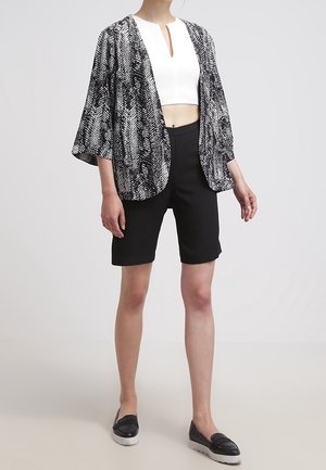 Kvinde iført sort og hvidt mønstret åben jakke, hvid cropped top, sorte knælange shorts og sorte slip-on sko.