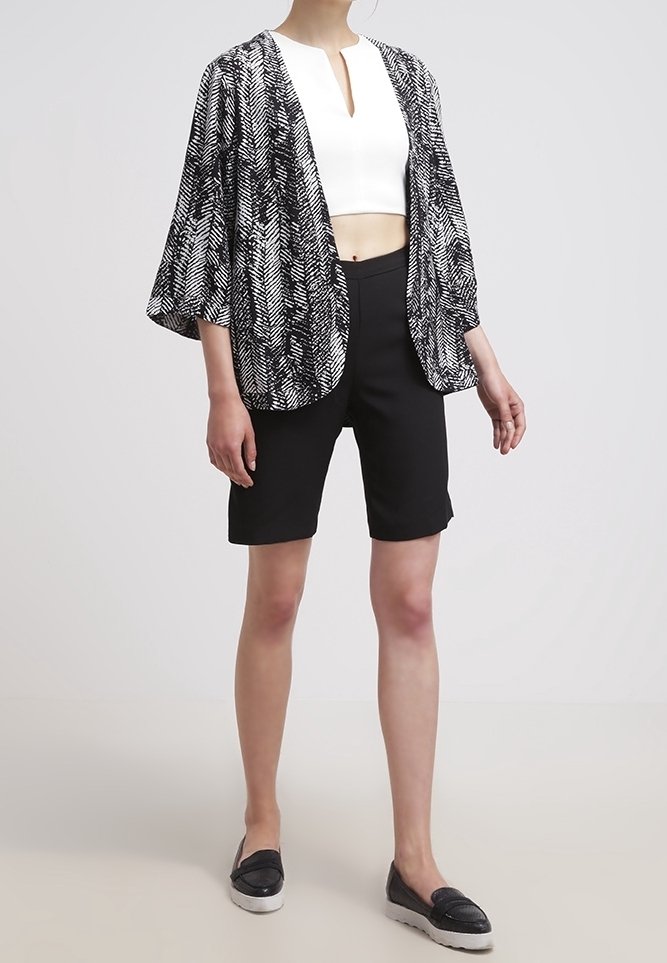 Femme portant une veste ouverte à motif noir et blanc, un top court blanc, un short noir au genou et des chaussures noires sans lacets.