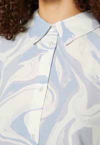 Gros plan d'une personne portant une chemise boutonnée avec un motif marbré bleu et blanc et un col pointu.