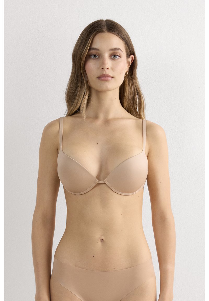 Donna con capelli lunghi in piedi contro una parete bianca, indossa reggiseno e biancheria intima color beige senza cuciture, guardando avanti con un’espressione neutra.
