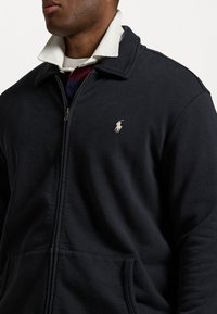 Man i en svart jacka med dragkedja och en liten broderad polo-spelare i vitt över en vit skjorta med krage.