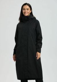 Kobieta uśmiechająca się, ubrana w długi czarny płaszcz przeciwdeszczowy z kapturem, zapinany z przodu na zamek i z bocznymi kieszeniami, stojąca na tle jednolitego jasnoszarego tła.