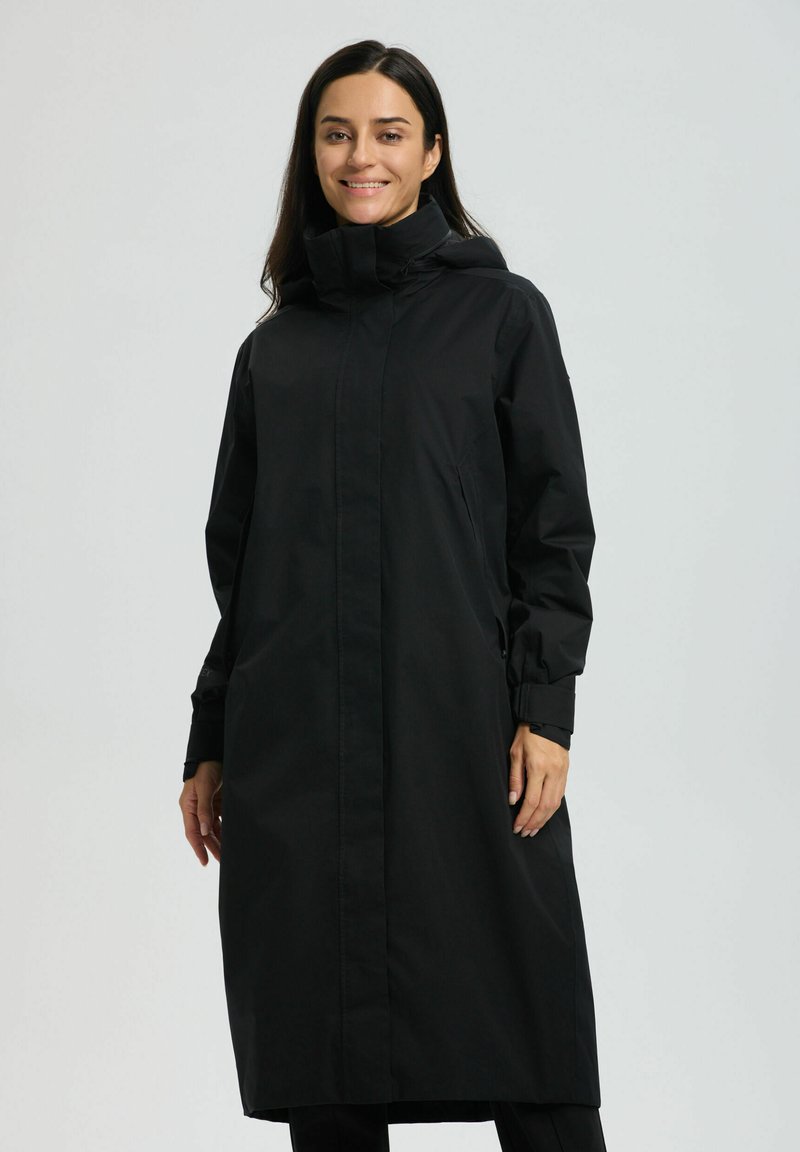 Kobieta uśmiechająca się, ubrana w długi czarny płaszcz przeciwdeszczowy z kapturem, zapinany z przodu na zamek i z bocznymi kieszeniami, stojąca na tle jednolitego jasnoszarego tła.