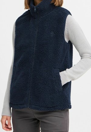 Femme portant un gilet en polaire bleu foncé zippé sur un pull gris clair à manches longues, une main dans la poche du gilet.