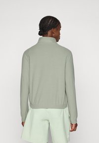 Light green pullover with a high collar, ribbed hem, and textured fabric. Worn over light green shorts, showing a relaxed fit and minimal design.