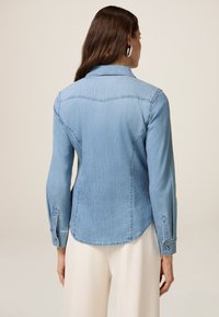 Cămașă din denim de un albastru deschis, cu guler cu nasturi, mâneci lungi și tiv curbat. Prezintă coasere tonale și un design clasic.