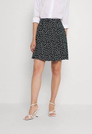 Vrouw die een zwarte A-lijn rok met witte polka dots draagt, een wit lang mouw shirt met opgerolde mouwen en witte sandalen met hakken, staand tegen een effen achtergrond.