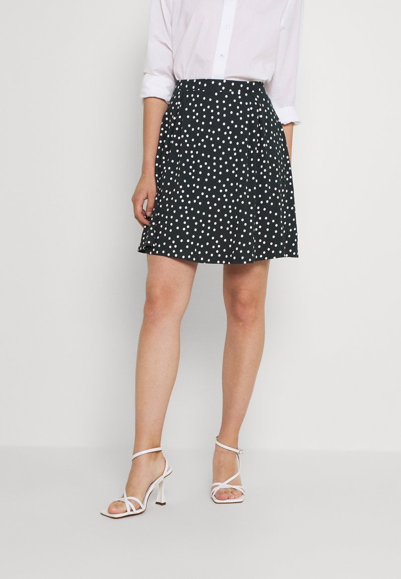 Femme portant une jupe noire trapèze à pois blancs, une chemise blanche à manches longues retroussées, et des sandales blanches à talons, posant devant un fond uni.