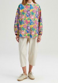 Osoba w luźnych, krótkich beżowych spodniach, beżowych butach sportowych i żółtej koszuli z guzikami w jaskrawy kwiecisty wzór w odcieniach fioletu, niebieskiego i różowego.