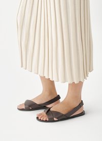 Kāji valkā tumši brūnas sandales ar krusteniskām siksniņām, kombinētas ar garu krēmkrāsas plīsētu svārku.