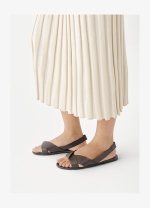 Stopy w ciemnobrązowych sandałach na obcasie z krzyżującymi się paskami, w zestawieniu z długą kremową plisowaną spódnicą.