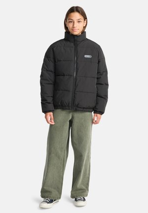 Giacca nera imbottita con colletto alto, dotata di chiusura con zip e una piccola etichetta con il logo. Abbinata a pantaloni verdi larghi e sneakers.