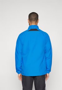 Modra jakna iz gladkega blaga, z visokim ovratnikom in črnimi poudarki na ramenih in hrbtu. Elastični manšeti in sproščen kroj.