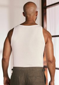 Weißes, geripptes Tanktop mit breiten Trägern, kombiniert mit olivgrün gestreiften Hosen. Der Hintergrund zeigt sanfte, neutrale Farben und zarte Texturen.