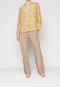 Femme portant un chemisier à manches longues jaune à motifs, un pantalon large beige et des chaussures beiges à talons ouverts, debout devant un fond blanc.